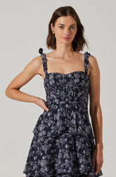 A woman is wearing a sleeveless dress with shoulder straps tied into small bows. The dress features a dark background with a gray floral pattern and a tiered, ruffled skirt. The fabric appears lightweight and textured.
